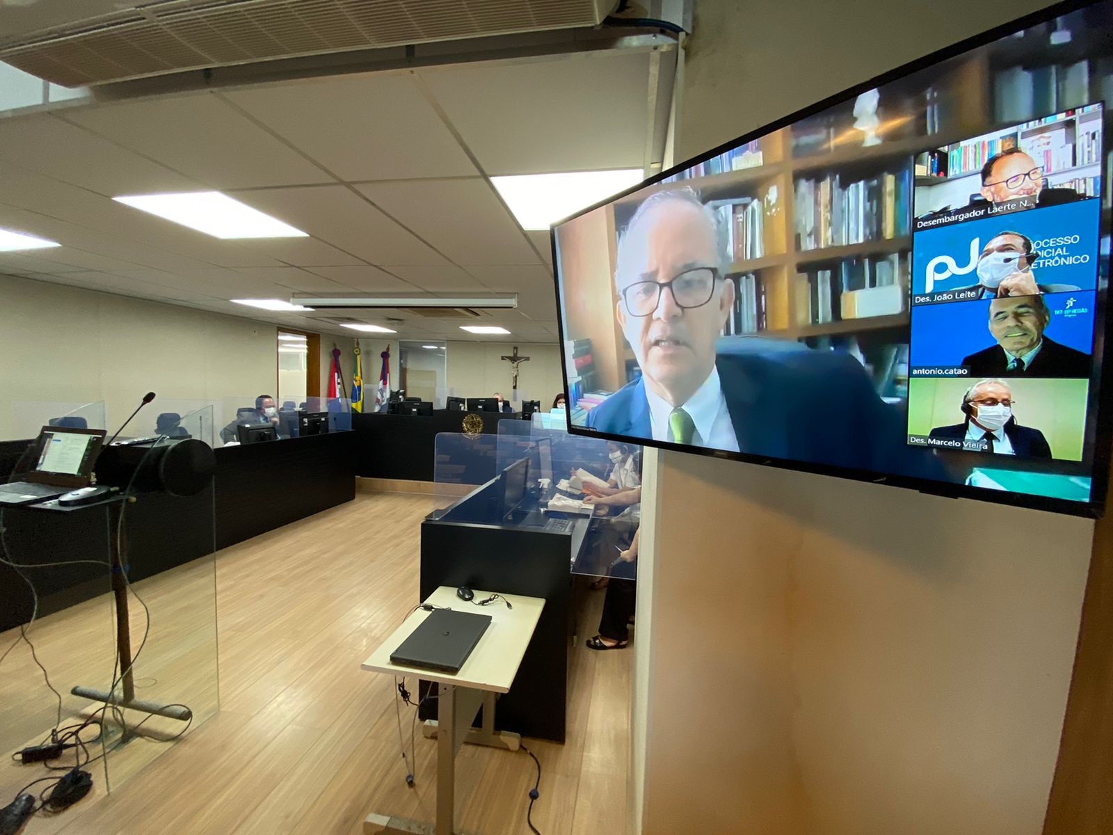 Tela de televisão fixada na parede da sala de sessões mostra os rostos de quatro desembargadores no ambiente da plataforma zoom, com a bancada do plenario ao fundo