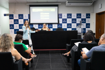 Mulheres e homens sentados, assistindo  à uma mulher de blazer azul, em pé, falar