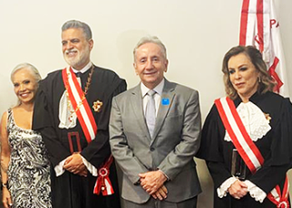Um homem alto, com cabelo, barba e bigodes brancos usando uma toga com faixa vermelha, uma mulher de vestido preto e branco, morena de cabelos longos e brancos, um homem um pouco mais baixo que o outro, de terno cinza, gravata cinza, blusa branca, cabelos brancos e sem barba e sem bigode e uma mulher loira, de pele branca usando toga e faixa vermelha
