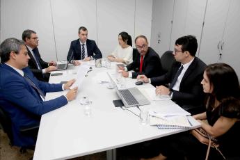 Reunião em volta de uma mesa retangular branca, com 5 homens brancos de terno e duas mulheres brancas