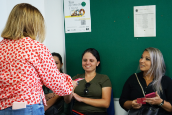 Mulher de blusa vermelha e branca entregando filpeta para três mulheres sentadas trajando roupas escuras.