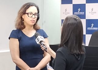 Mulher de pele morena, de cabelos longos e pretos, usando óculos de armação preta e um vestido azul marinho concedendo entrevista a uma repórter de cabelos pretos.