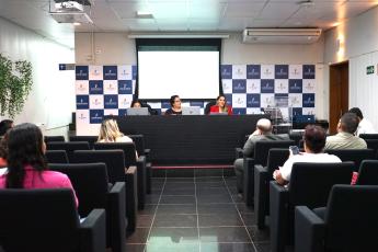 Mesa de auditório composta com três pessoas  de pele clara, sendo duas delas de roupa escura e uma de blusa vermelhas falando para divresas pessoas na plateia