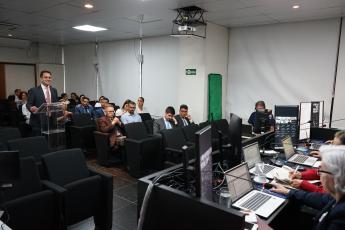 Auditório com várias pessoas em frente a uma bancada. Há um homem de peleclara falando para mulheres e homens em uma bancada