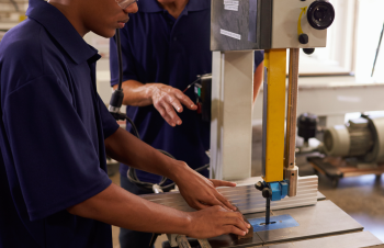 Duas pessoas com peles claras vestidas de roupas na cor azul manuseando uma máquina de cor bege e amarela