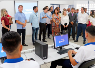 Homens e mulheres de várias etnias de pé trajando roupas em tons claros e escuros ouvindo homem falando, de frente a dois homens jovens sentados a frente de computadores, vestidos de camisas nas cores branca e azul.