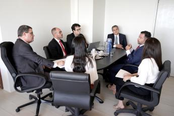 Foto da mesa com juízes e servidores vestidos de ternos pretos e duas mulheres de cabelos longos e pretos  sentadas  de costas com blusas branca