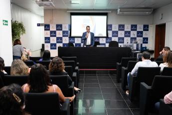 Homem de terno azul escuro e camisa branca falando para público sentado em cadeiras pretas em um auditório.