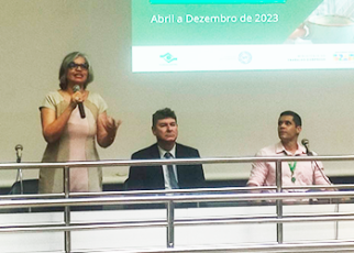 Mulher branca de vestido verde e rosa de pé falando. Há dois homens sentados, sendo um de terno escurp e outra de camisa rosa claro