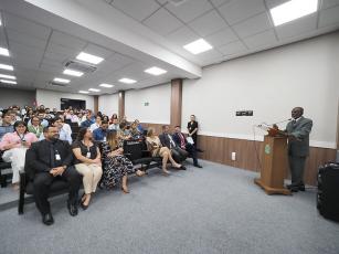 Foto em diagonal registrando várias mulheres e homens sentados em um auditório. Na frente das pessoas, em um púlpito, está o presidente do TRT-11, desembargador Audaliphal Hildebrando, um homem negro e de terno cinza. 