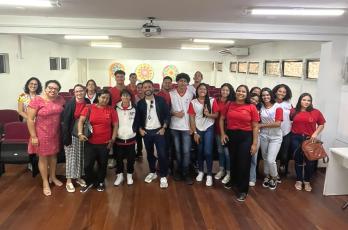 Foto com muitas pessoas, homens e mulheres, em pé. Piso de madeira e parede, ao fundo, na cor branca.