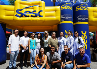 Grupo de mulheres e homen, alguns acocorados, em frente a uma tenda inflável amarela e azul
