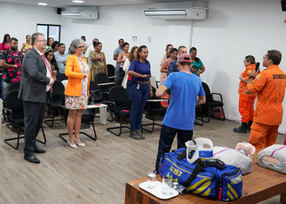 Vários homens e mulheres de pé prestando atenção a fala de dois bombeiros fardados de cor laranja e um homem de camiseta preta e calça preta.