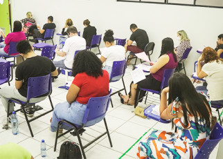 Vários homens e mulheres sentados em carteiras escolares de cor azul escrevendo.