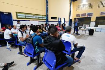 Homem de terno escuro e camisa azul falando para diversas alunos de uniformes brancos sentados em carteiras escolares na cor azul.