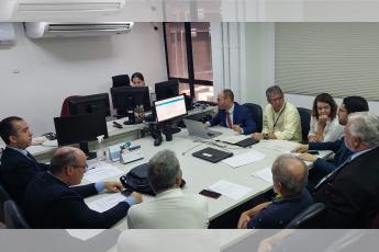 Foto de uma audiência de conciliação, com cinco advogados usando ternos escuros; um juiz usando terno azul-marinho, um servidor de camisa amarelo claro e uma advogada usando blusa branca