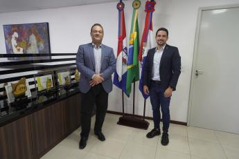 Dois homens de pé na frente de três bandeiras. Ele estão trajando ternos.