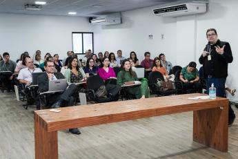 Sala de aula com dezenas de pessoas sentadas, olhando atentamente o professor , que está à frente, usando terno escuro e gesticulando com uma das mãos