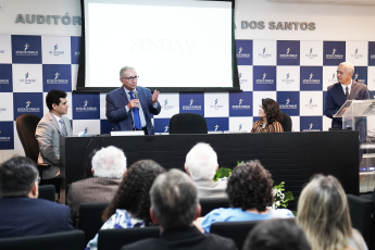 Homem de cabelos grisalhos e paletó azul claro discursando ao centro da mesa, à esquerda, homem sentado de cabelo preto e paletó cinza claro; à direita, mulher sentada de cabelo preto e blusa mostarda e listras pretas e um homem de paletó preto em pé no púlpito