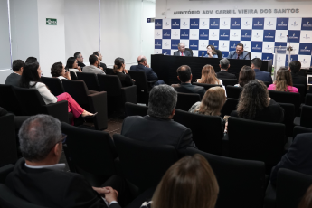 Auditório com homens e mulheres reunidos . Há dois homens de ternos escuros e uma mulher entre eles. Ela veste roupa azul e está falando.