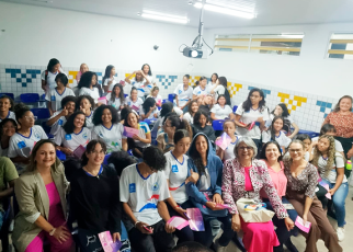 Plateia com vários alunos uniformizados de camisetas brncas e calças jeans. Há  várias mulheres com vestidos de diversas cores.