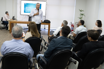 Homem branco de camisa branca com microfone na mão falando para um grupo de pessoas sentadas