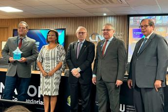 Desembargador João Leite de Arruda Alencar, à esquerda, com terno cinza escuro e gravata vermelha, segurando um troféu de acrílico azul, acompanhado, ao seu lado esquerdo, por uma mulher usando vestido branco gelo com flores cáquis e por três homens, um vestindo terno preto, e dois usando ternos cinzas escuros.