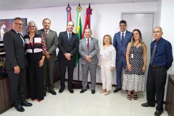 Grupo de homens e mulheres posando para fotos. Ele estão em uma sala branca, na qual há três bandeiras, sendo a do Brasil localizada ao centro.