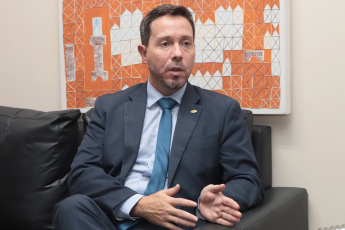 Homem branco de meia idade, vestido de terno escuro. Elé está sentado em um sofá também escuro. Há um quadro nas cores laranja e branco na parede detrás dele.   