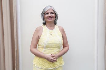 Mulher de pele morena, cabelos curtos e brancos, de vestido amarelo em pé de braços cruzados