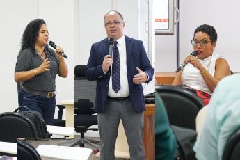 À esquerda, mulher morena, baixa, de cabelo preto em pé com um microfone na mão esquerda; ao centro, um homem  em pé, calvo, moreno, de óculos, paletó azul-marinho, gravata azul marinho e blusa branca; à direita, uma mulher morena, sentada, de cabelo preto e curto, usando blusa branca 