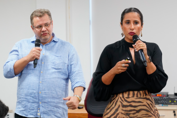 Homem branco de óculos com camisa de manga comprida azul. Há uma mulher de pele clara vestida de blusa de manga comprida preta e saia bege com listras horizontais na cor preta. Os dois têm seguram microfones.