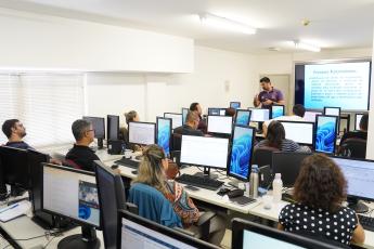 Vários servidores sentados, de costas,  à frente de seus computadores; à frente, homem de altura mediana, cabelo preto, em pé lecionando a aula, usando camisa de cor roxa.