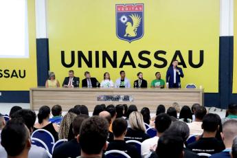 Público sentado assitindo palestra. Há uma mesa grande composta de várias pessoas. Há um homem de terno escuro de pé. Ele está falando ao microfone.
