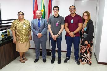 Três homens e duas mulheres posando para uma foto. Eles estão de pé. As pessoas estão na frete das bandeiras do Brasil, de Alagoas e do TRT-19.