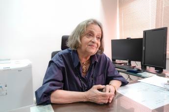 Mulher idoso sentada à mesa; ela tem pelo morena e cabelo grisalho e usa uma blusa azul marinho; ao lado esquerdo dela, a imagem de dois monitores, um em formato horizontal e outro vertical