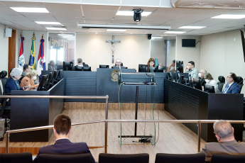 Sala de audiência com homens e mulheres sentados atrás de bancadas com computadores e um púlpito transparente no meio do palco