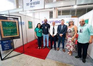 Grupo de homens e mulheres em pé dentro de um stand com a placa TRT-19