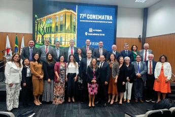 Grupo de pessoas, homens e mulheres, todos de pé, trajando roupas sociais, tendo ao fundo um painel com a inscrição 77º Conematra