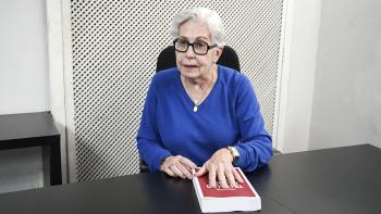 Foto da desembargadora Anne Inojosa. Ela é uma mulher branca de cabelo grisalho, usa óculos pretos e blusa azul. Ela está sentada em uma cadeira preta atrás de um birô preto com a mão esquerda sobre um livro vermelho.