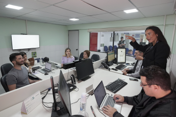 Sala de audiência. Há u,ma homem sentado em frente a um notebook. Ele está digitando. Há quatro pessoas sentadas olhando uma mulher de cabelos longos eescuros utilizando gestual da linguagem de libras.