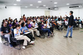 Home moreno, alto, magro, de blazer preto, em pé; frente dele, vários estudantes sentados escutando a sua palestra