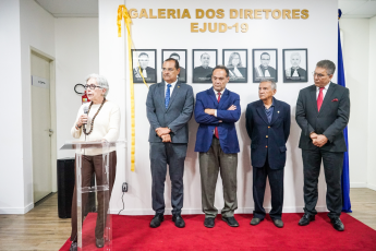 À esquerda, em pé, mulher branca de cabelo grisalho discursando no púlpito com o microfone na mão, à direita, quatro homens em pé usando paletós escuros