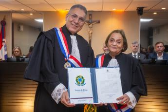À esquerda, homem alto de cabelo  grisalhos e de toda preta; à direita, mulher morena baixa e idosa também de toga; ambos estão segurando o certificado branco grande.