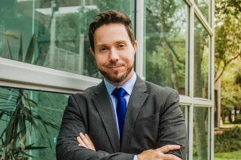 Há um homem branco, com cabelo e barba claros. Ele está vestido com um terno escuro, camisa azul-clara e gravata azul-escura. De braços cruzados, ele sorri.