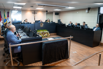 Na foto, vários homens de paletó sentados do lado direito e do lado esquerdo atrás de uma bancada preta, ao centro dessa bancada, um homem de paletó azul e ao lodo esquerdo dele uma mulher de vestido verde e outra mulher de roupa bege.