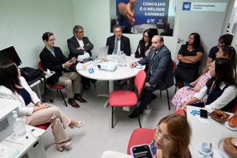 Vá rias pessoas reunidas em uma sala. Há uma mesa rendonda no centro da sala.