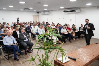 Um auditório com várias pessoas sentadas. Elas assistem um palestra feita por um homem que veste terno escuro.