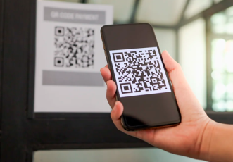 Há uma mão de pessoa branca com o celular na mão com um QR Code na tela.