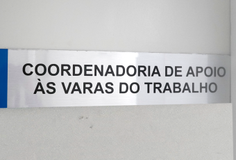 Pla de metal cinza com letras pretas. Há escrito Coordenadoria de Apoio às Varas do Trabalho.